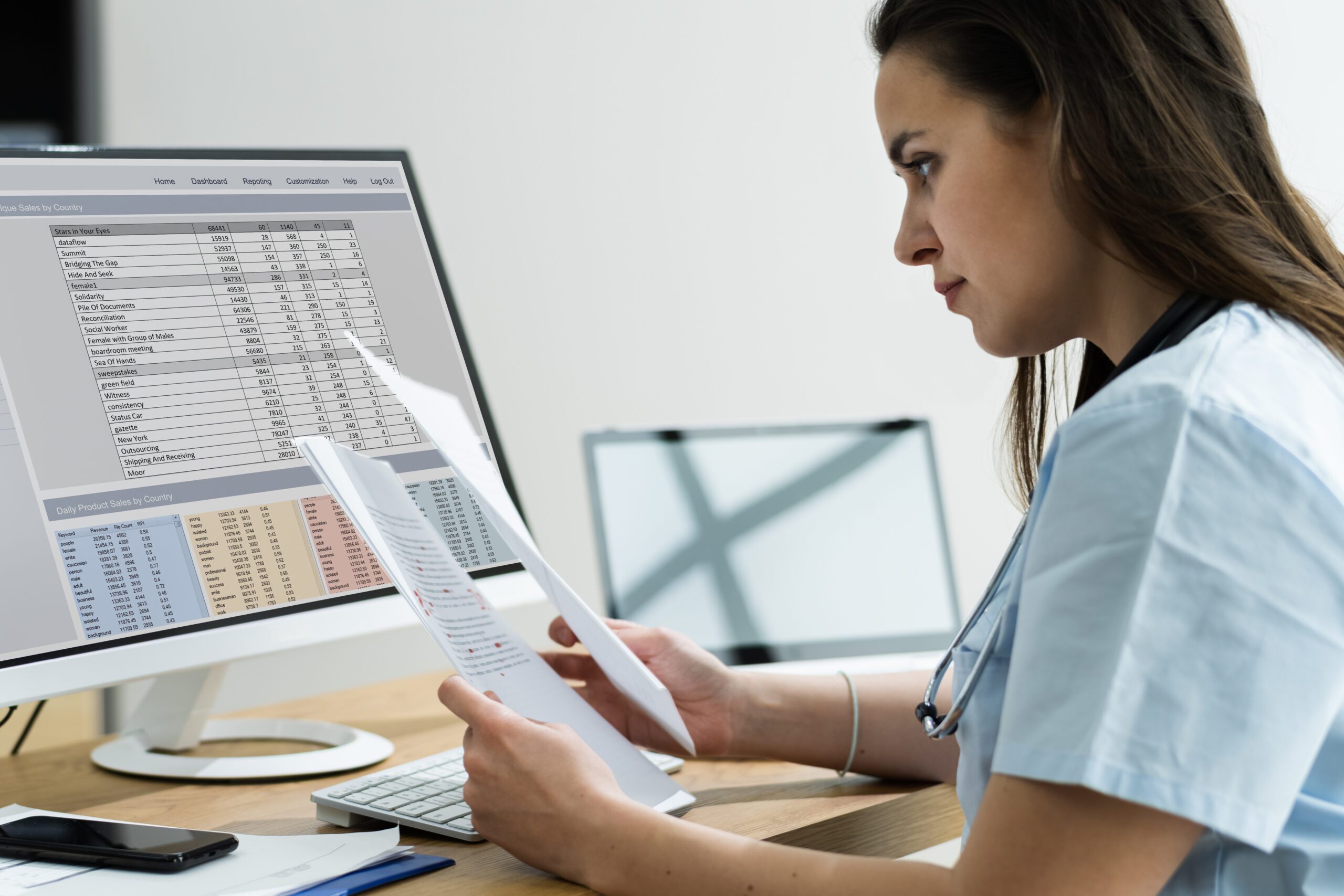 Female doctor examining papers in front of a desktop computer with spreadsheets on the screen